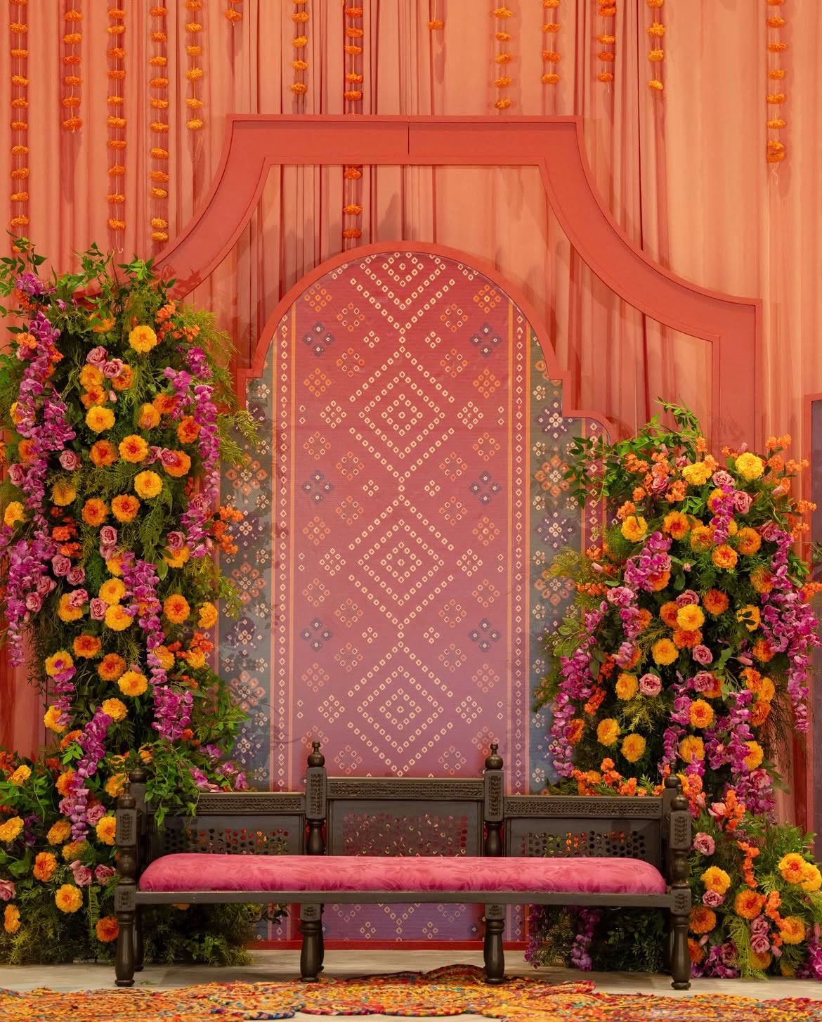 Warm peach and magenta scalloped arch backdrop with cascading marigolds and floral towers for mehendi rituals