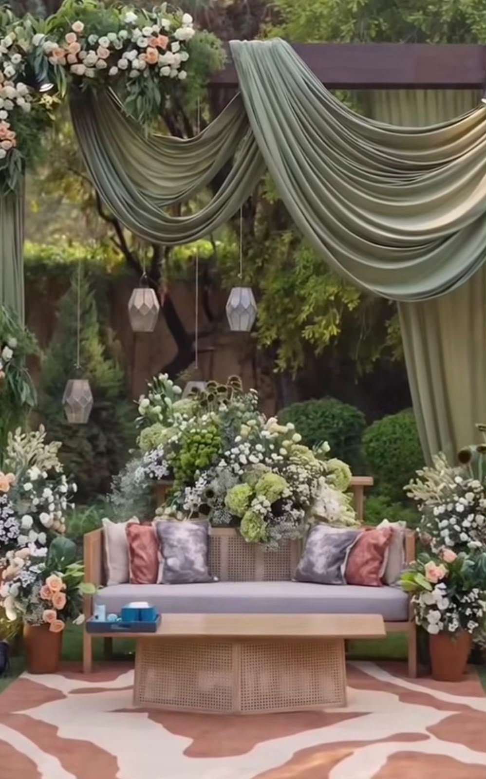 Sage green draped garden canopy with hanging geometric lanterns, lush botanical backdrop, and comfortable guest lounge