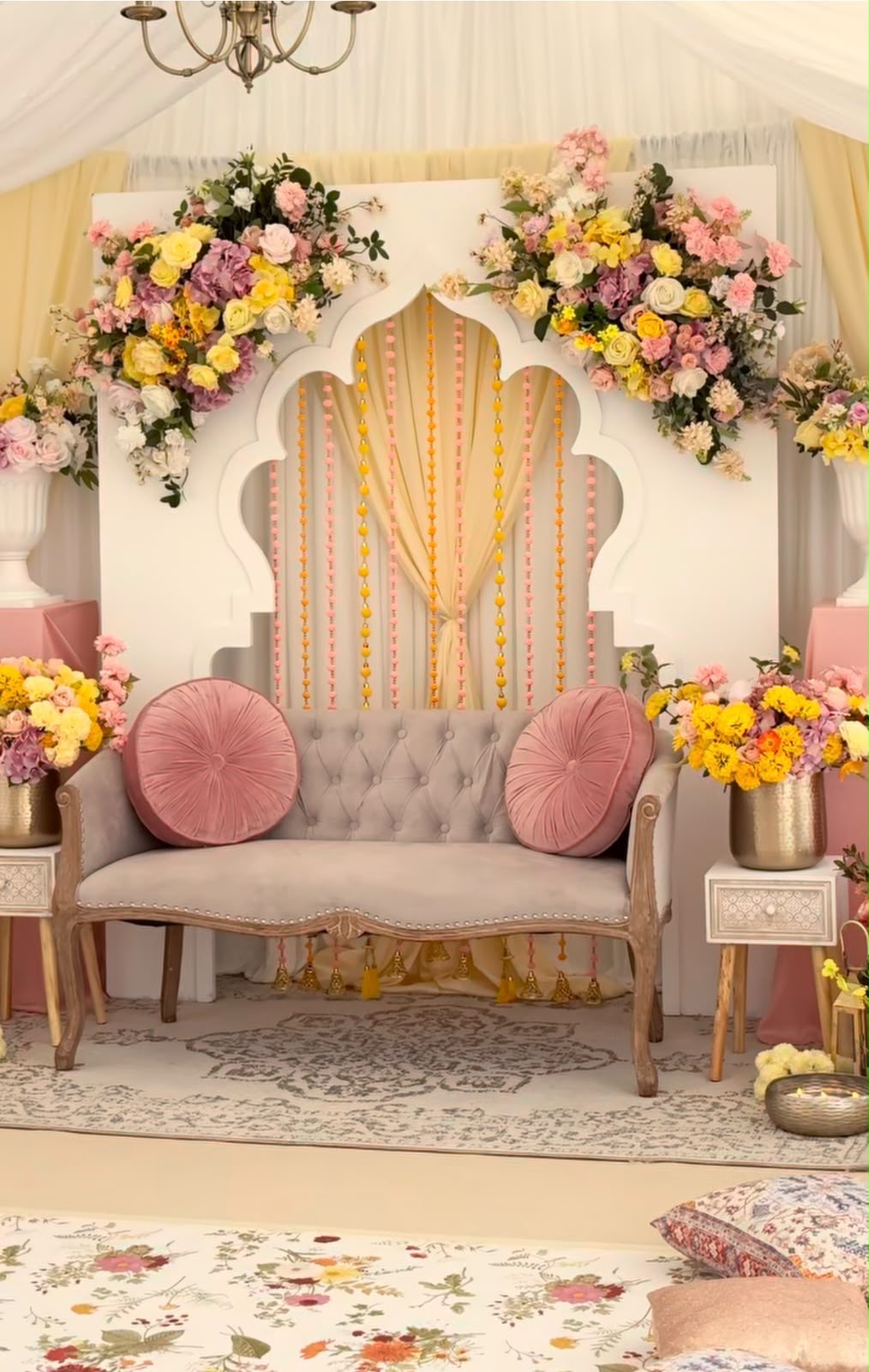 White Mughal arch mehendi stage with yellow marigold tassels, tufted velvet sofa, and blooming golden urn arrangements