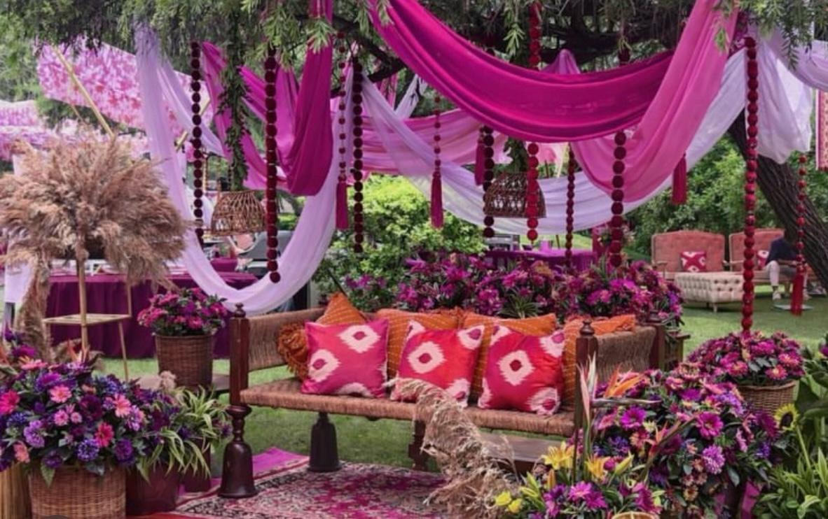 Bohemian open-air lawn mehendi setting under lush trees with magenta chiffon drapes and vibrant flower baskets
